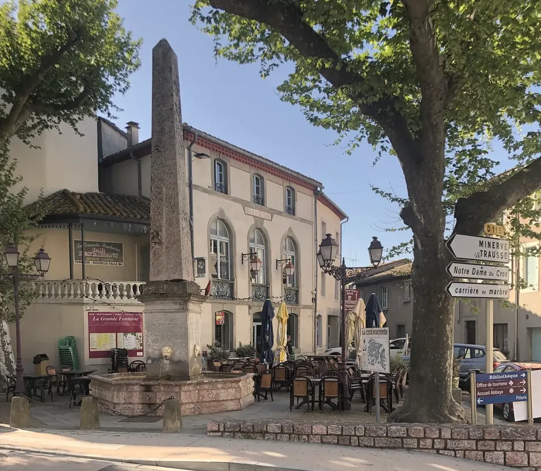 La terrasse de La Grande Fontaine sur la place de Caunes-Minervois : la fontaine et son obélisque de marbre au premier plan, la façade du restaurant et les tables sous les platanes centenaires.